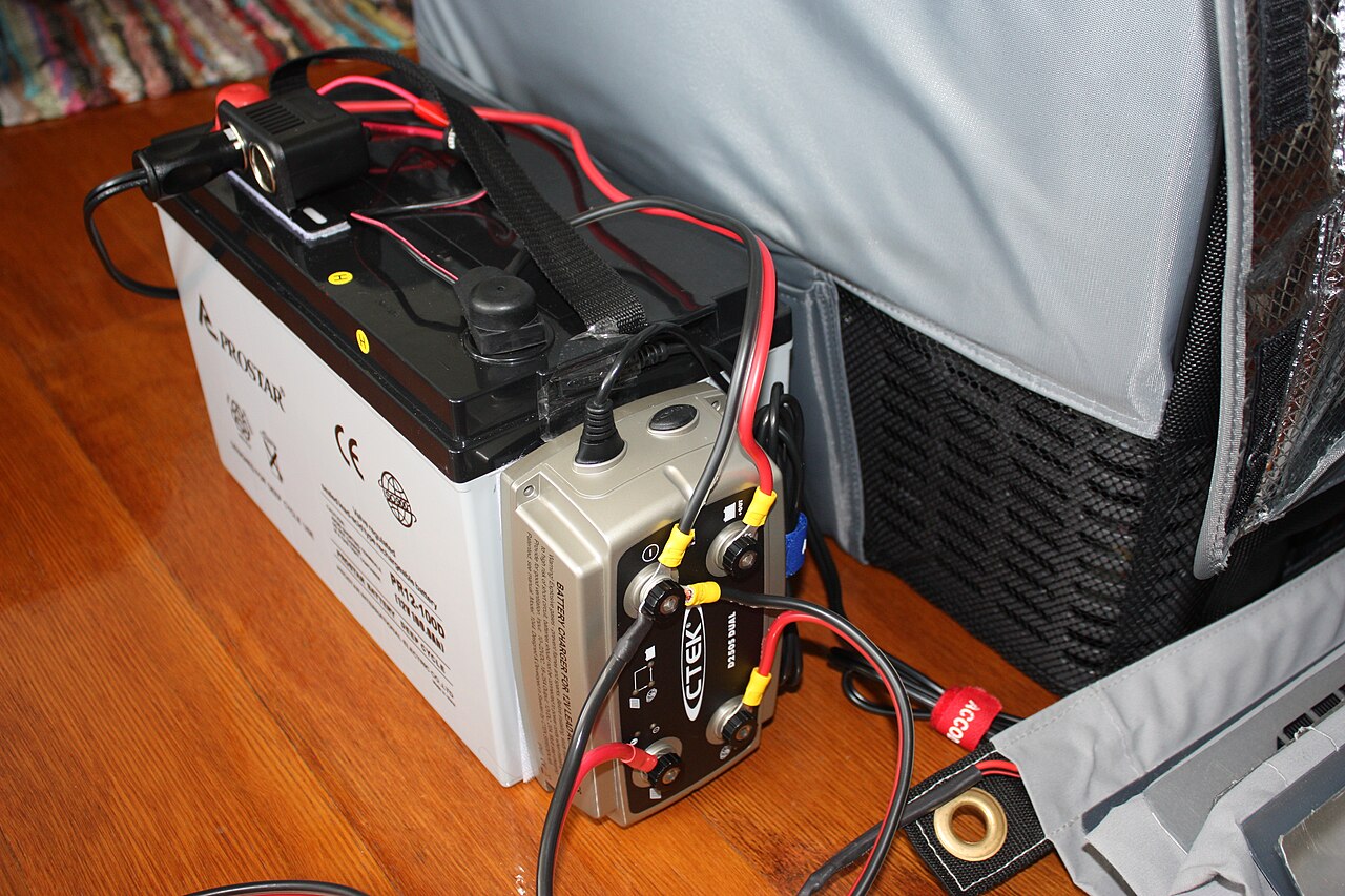 Ein DC-DC-Ladegerät mit angeschlossenen roten und schwarzen Kabeln mit Ringkabelschuhen, seitlich an einer 12-Volt-Zusatzbatterie befestigt.